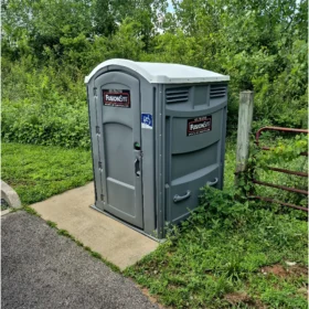 A gray portable restroom with a wheelchair-accessible symbol is situated on a concrete pad surrounded by lush greenery and a wooden fence in a rural outdoor setting.