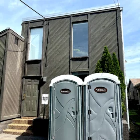 Two gray portable restrooms are positioned side by side on a paved area in front of a dark brown building with large windows and a green door on a sunny day.