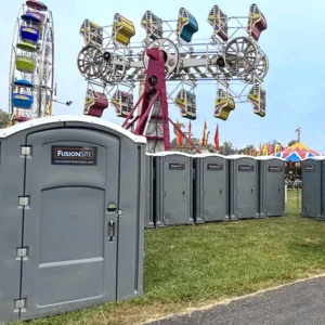 A row of gray portable restrooms is set up on grass beside a paved walkway, with colorful carnival rides and tents visible in the background on an overcast day.