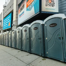 A row of gray portable restrooms is lined up against a building's exterior, with large digital billboards displaying advertisements above them on a sunny day.