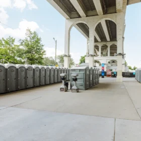 A row of gray portable restrooms is positioned along a concrete pathway beneath a large overpass, with trees and a modern building visible in the background on a partly cloudy day.