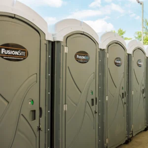 A row of gray portable restrooms is lined up along a concrete walkway beneath a large overpass, with parked vehicles and a construction site visible in the background on a cloudy day.