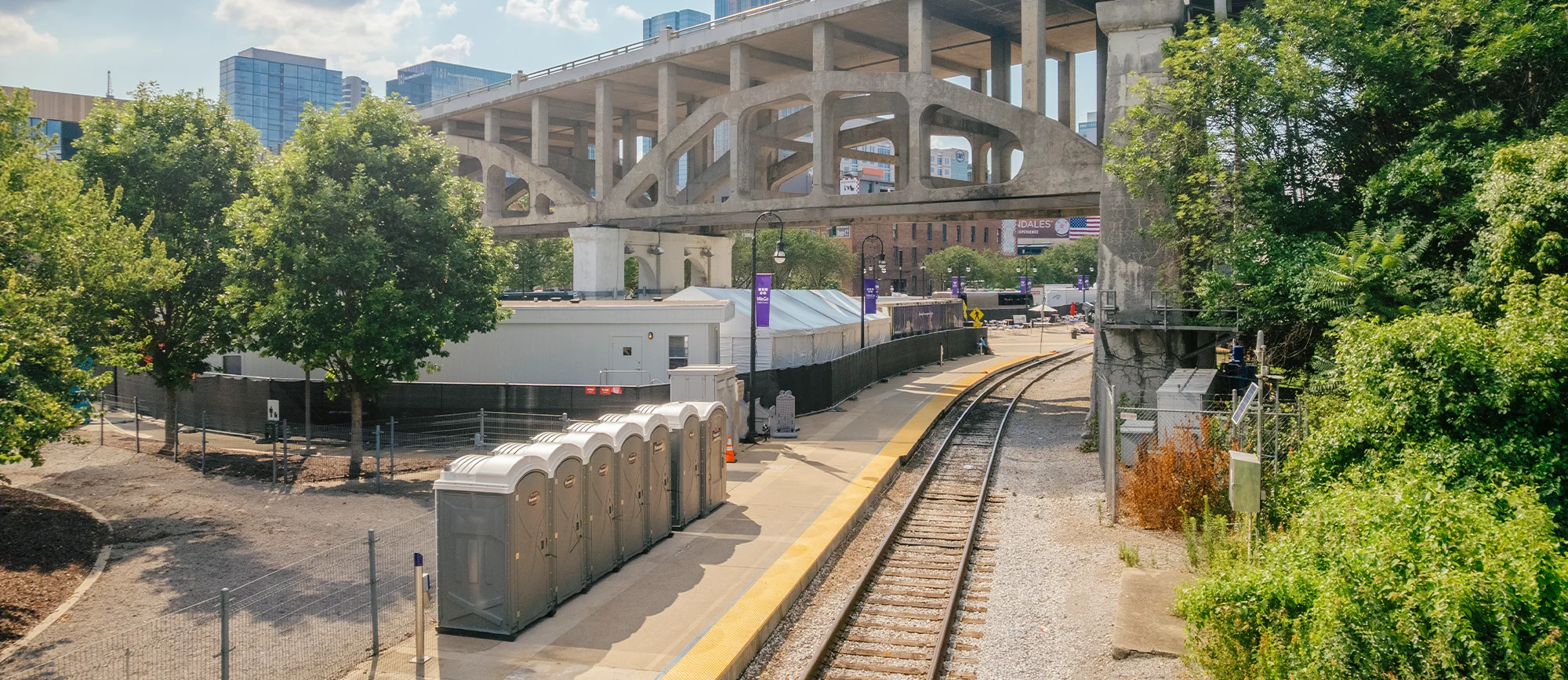 A row of gray portable restrooms is positioned along a concrete platform beside a railway track, with a large concrete overpass and city buildings visible in the background under a partly cloudy sky.