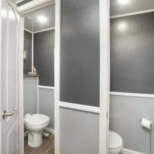 Interior of a gray restroom trailer featuring two private toilet stalls with white trim, wood-style flooring, and soft overhead lighting for a clean, modern appearance.