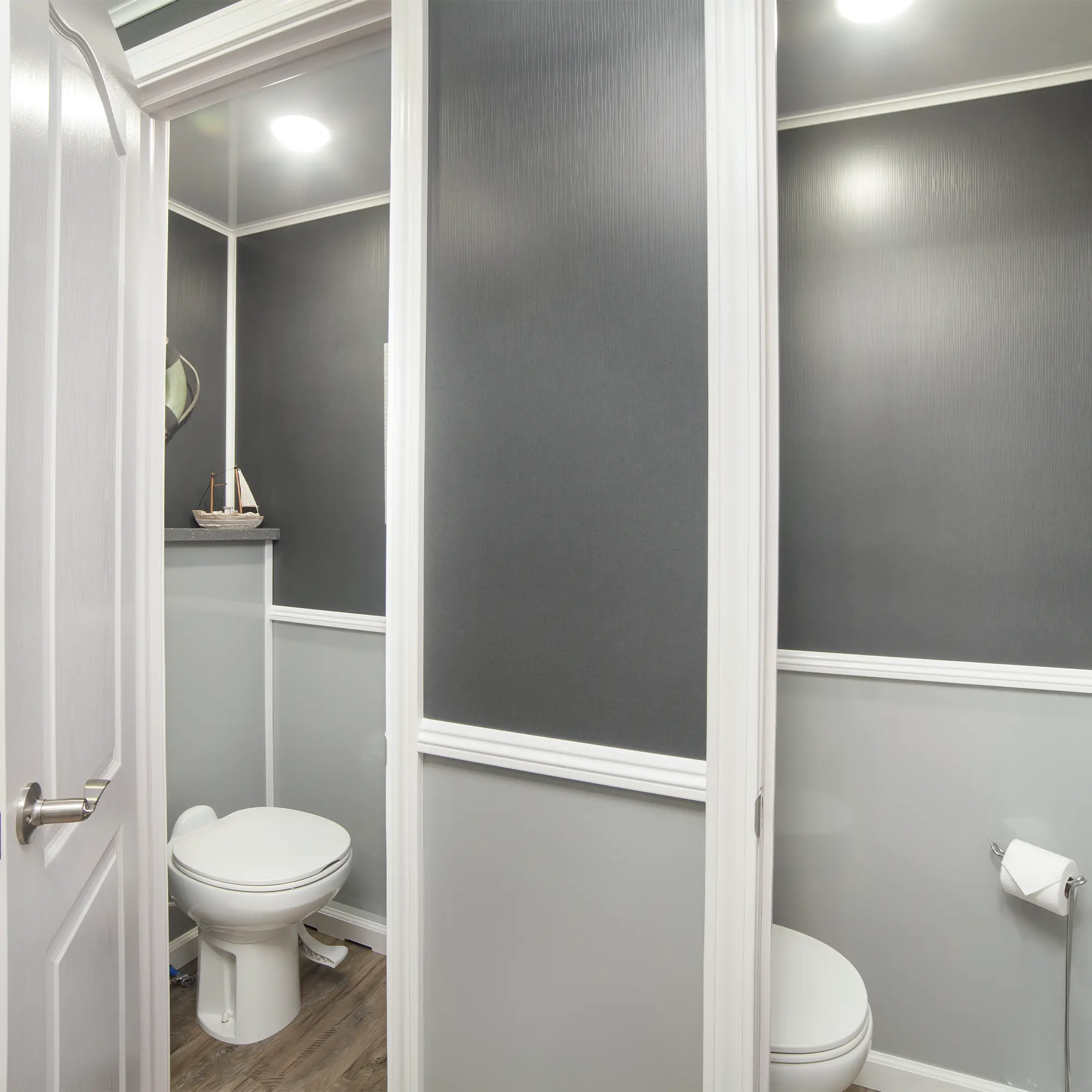 Interior of a gray restroom trailer featuring two private toilet stalls with white trim, wood-style flooring, and soft overhead lighting for a clean, modern appearance.