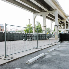 A row of green portable restrooms is positioned behind a chain-link fence in a parking lot under a concrete overpass, with a modern building visible in the background.