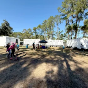 A group of people walks across a grassy area with several portable restrooms and service trailers lined up under tall trees on a clear day.