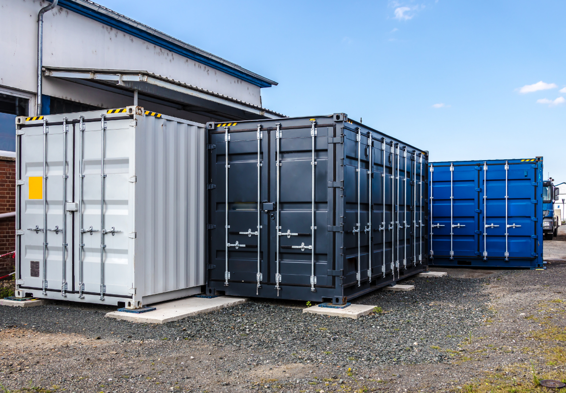 A row of three shipping containers in varying colors is positioned on a gravel surface beside a brick building, with a clear blue sky overhead.