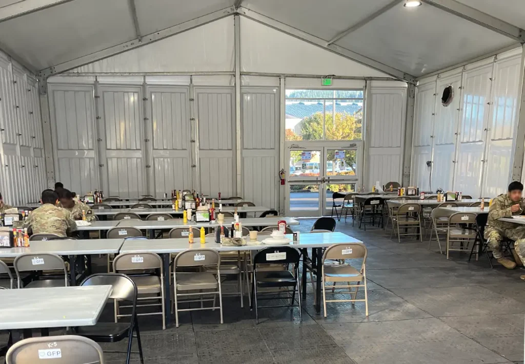 An indoor dining tent with several rows of folding tables and chairs, featuring condiment bottles, is partially filled with individuals seated and eating, while a large entrance opens to an outdoor view.