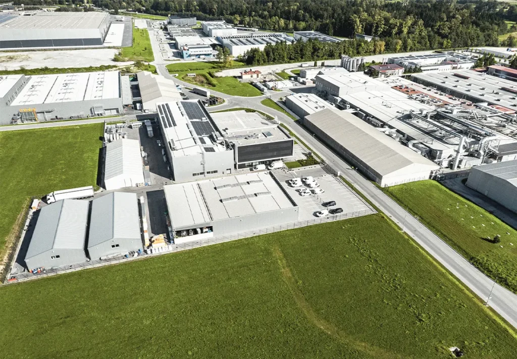 An aerial view of a large industrial complex featuring several gray warehouses, parking lots, and green grass areas, with a road running alongside the property under a clear blue sky.