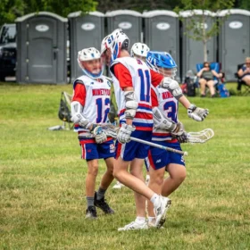 A group of young lacrosse players in red and blue uniforms celebrate on a grassy field, with several portable restrooms visible in the background and spectators seated under trees.