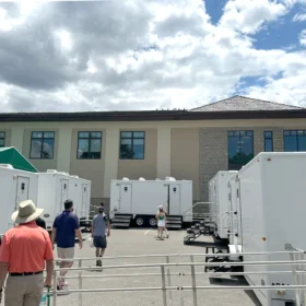 Multiple white restroom trailers are lined up outside a large building at a golf tournament, with attendees walking between units under a partly cloudy sky.