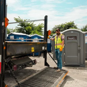 A man wearing a safety vest and gloves stands on a loading ramp beside a truck, with several portable restrooms lined up in the background under a blue sky.