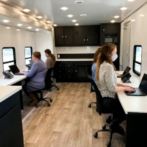 Inside a mobile office trailer, three masked women are seated at desks with laptops, working under bright lights and near windows, with dark cabinetry and a neutral-colored floor.