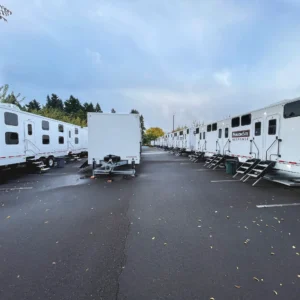 A row of white mobile restroom trailers is parked on an asphalt lot, lined up beside a large white storage trailer under a cloudy sky, with scattered leaves on the ground.