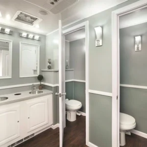 A clean, modern restroom trailer interior features two sinks with mirrors, two private toilet stalls, and soft lighting against light green walls and dark wood flooring.