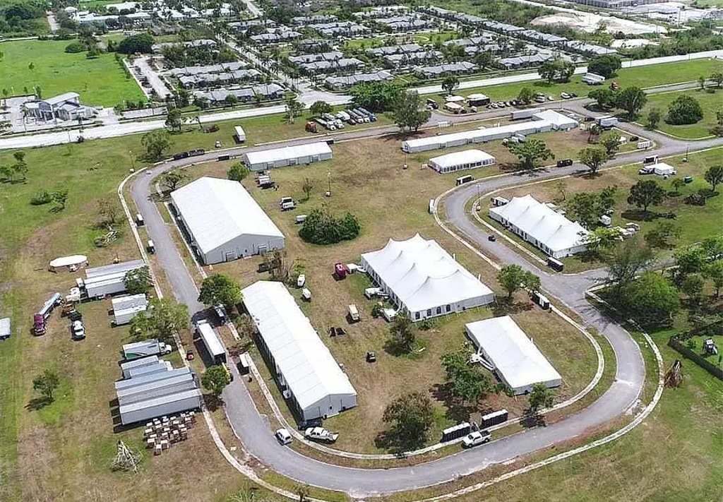 An aerial view of a large outdoor event site featuring several white tents, parked vehicles, and a winding road surrounded by green grass and trees on a sunny day.