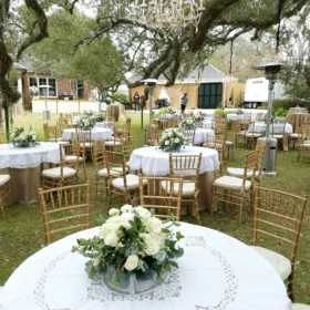 A beautifully arranged outdoor event space features round tables with white tablecloths and floral centerpieces, surrounded by gold chairs and patio heaters, with a restroom trailer visible in the background.