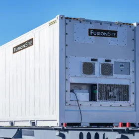A white portable refrigerated unit with ventilation and branding is positioned on a gravel lot, surrounded by trees and a clear blue sky in the background.