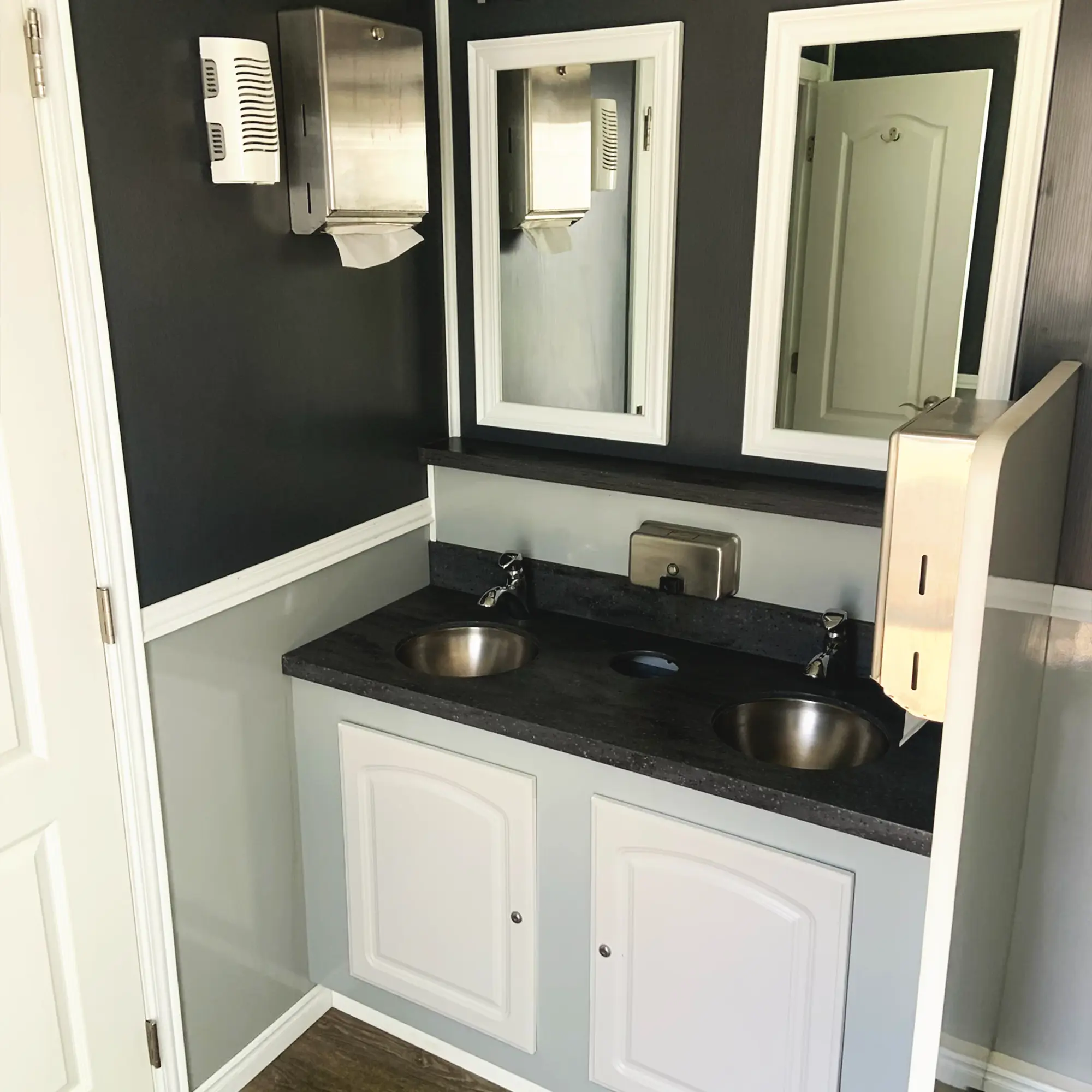 A clean, modern portable restroom interior features two stainless steel sinks, a large mirror, and a paper towel dispenser against a dark wall, with a white door leading outside.