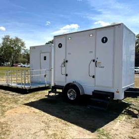 A white portable restroom trailer with two doors and an accessible ramp is parked on a grassy area under a blue sky, surrounded by trees and vehicles in the background.