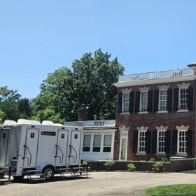 A white portable restroom trailer is parked on a paved area beside a large brick building with black shutters and tall columns, under a clear blue sky and surrounded by green trees.