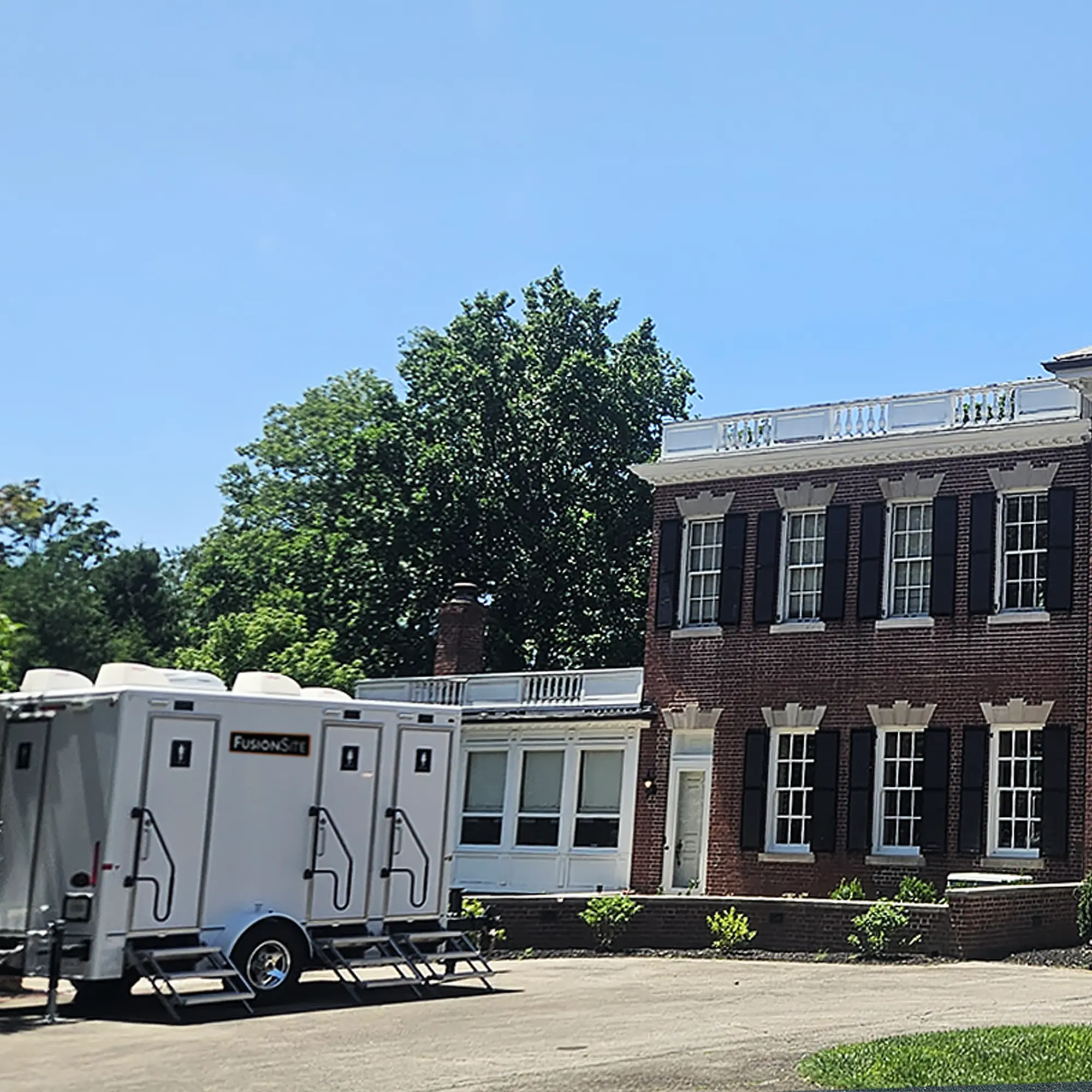 A white portable restroom trailer is parked on a paved area beside a large brick building with black shutters and tall columns, under a clear blue sky and surrounded by green trees.