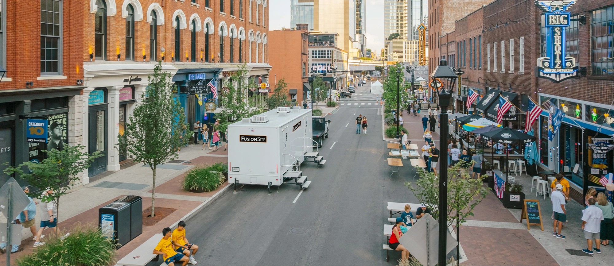 A white portable restroom trailer is parked on a busy street lined with shops and outdoor seating, surrounded by pedestrians and trees on a sunny day.