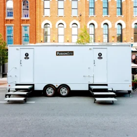A large white portable restroom trailer with two entry doors is parked on a paved lot surrounded by trees, with a car visible in the background on a sunny day.
