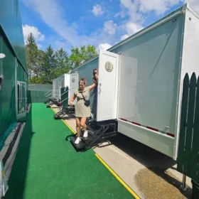 Two people are exiting a portable restroom trailer with steps, positioned on a green pathway beside a fenced area, under a bright blue sky with scattered clouds.