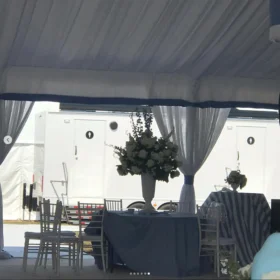 A white portable restroom trailer with a "Gotta Go" logo is positioned outside a tent, beside elegantly set tables and floral arrangements, under a canopy on a sunny day.