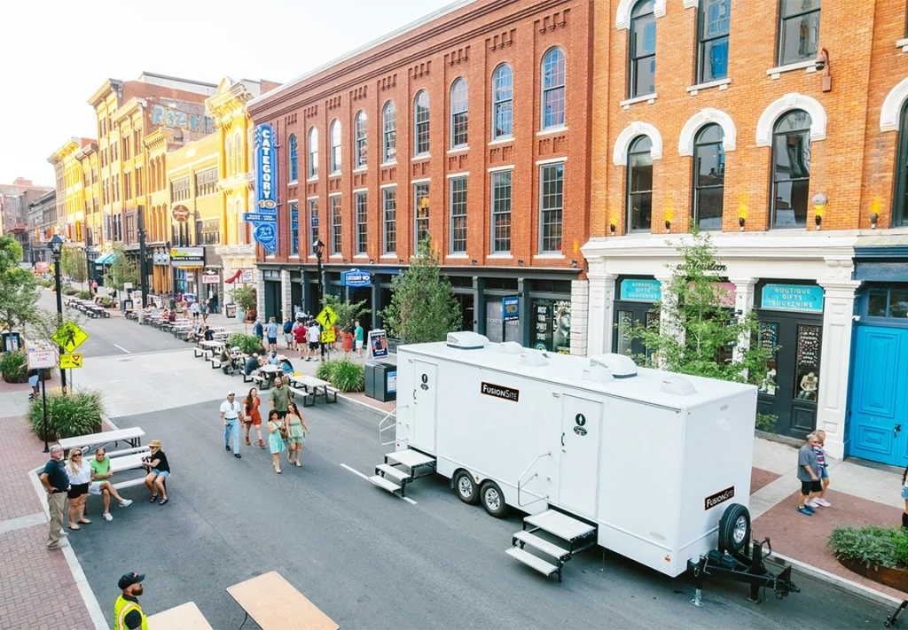 A white portable restroom trailer is parked along a bustling street lined with historic brick buildings, surrounded by people walking and outdoor seating on a sunny day.