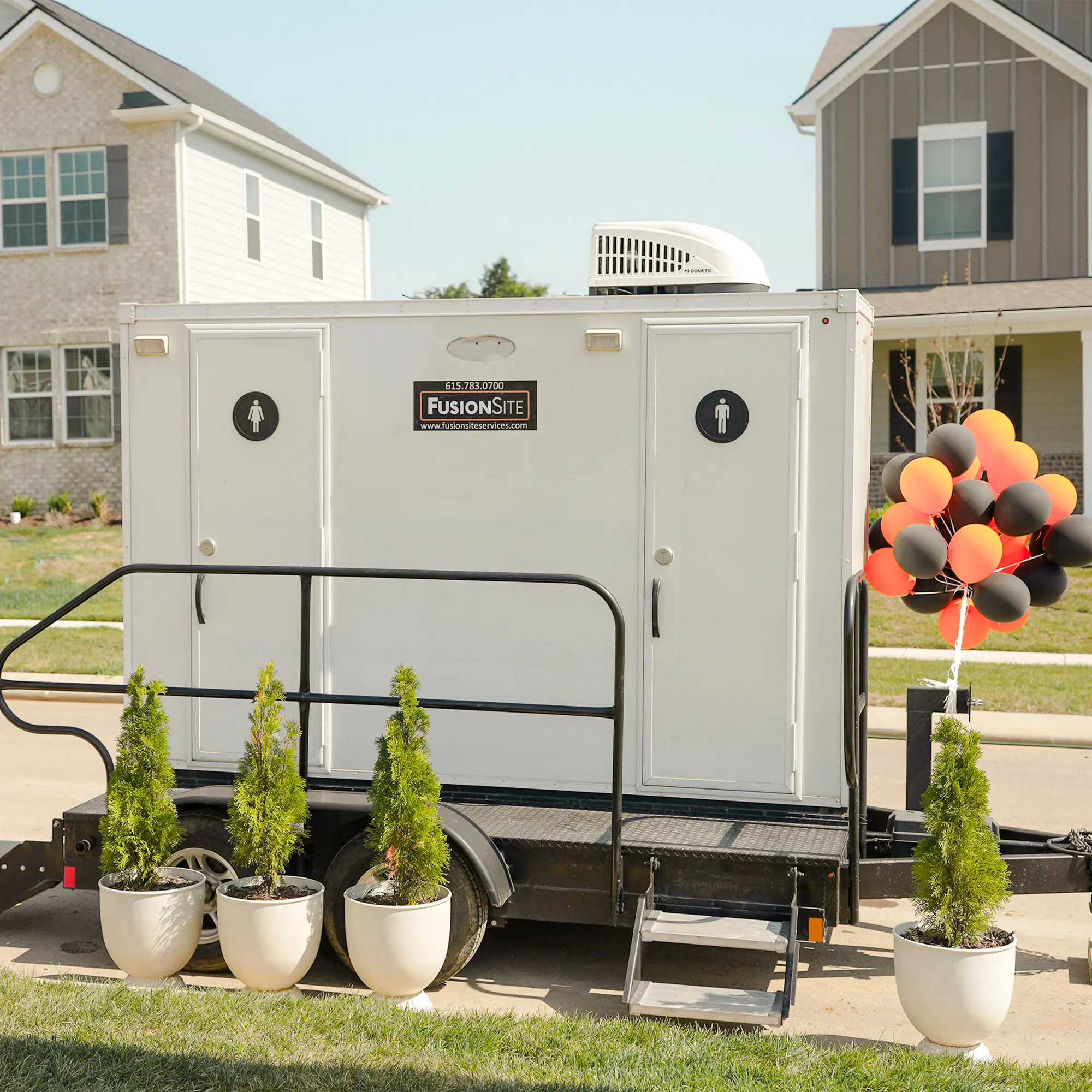 A portable restroom trailer with two entrances is parked on a paved driveway in front of a newly constructed residential home, surrounded by decorative potted plants and colorful balloons.