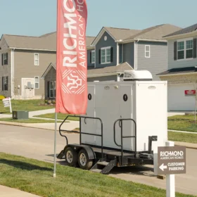 A portable restroom trailer with two doors is parked on a paved street in front of residential homes, flanked by red promotional flags and a customer parking sign on a clear day.