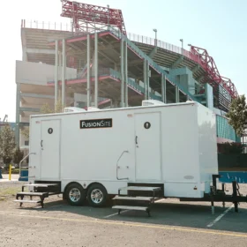 A white portable restroom trailer with two entrances is parked on a gravel lot next to a large stadium under construction, with trees and a blue barrier visible in the background.
