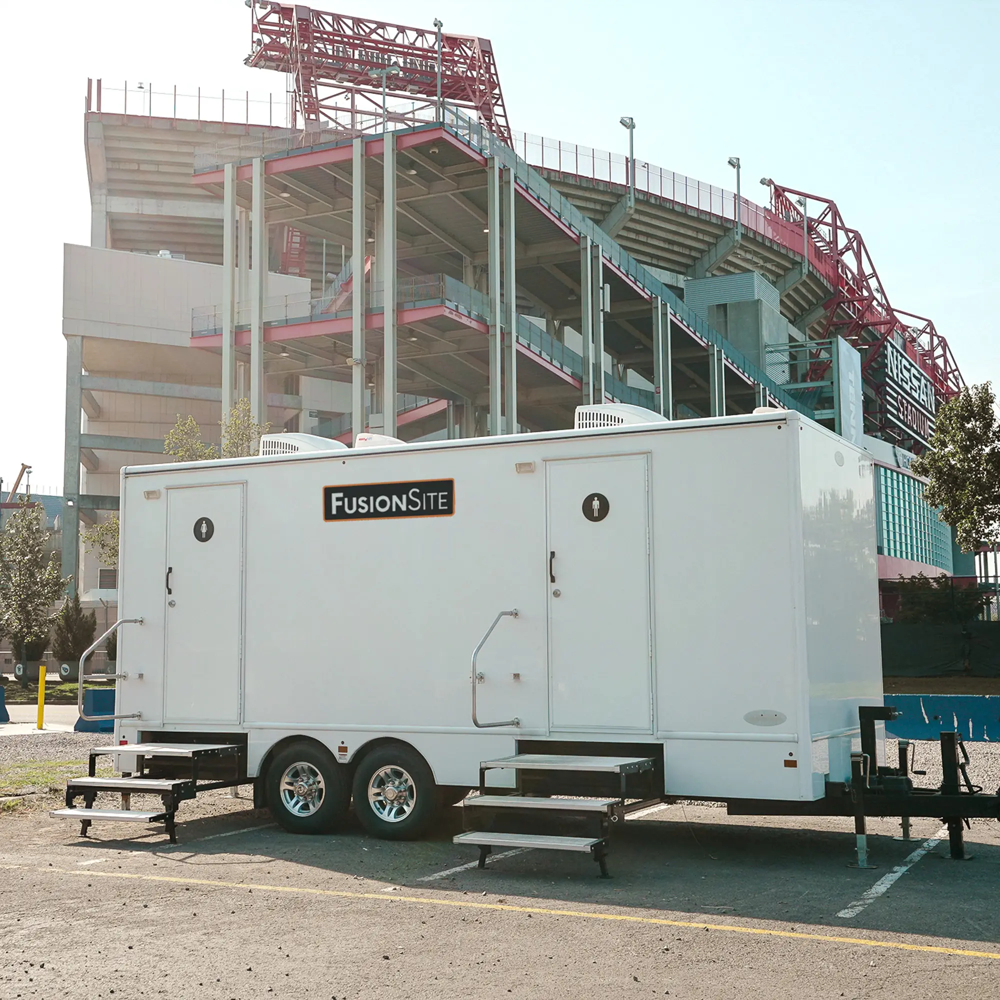 A white portable restroom trailer with two entrances is parked on a gravel lot next to a large stadium under construction, with trees and a blue barrier visible in the background.