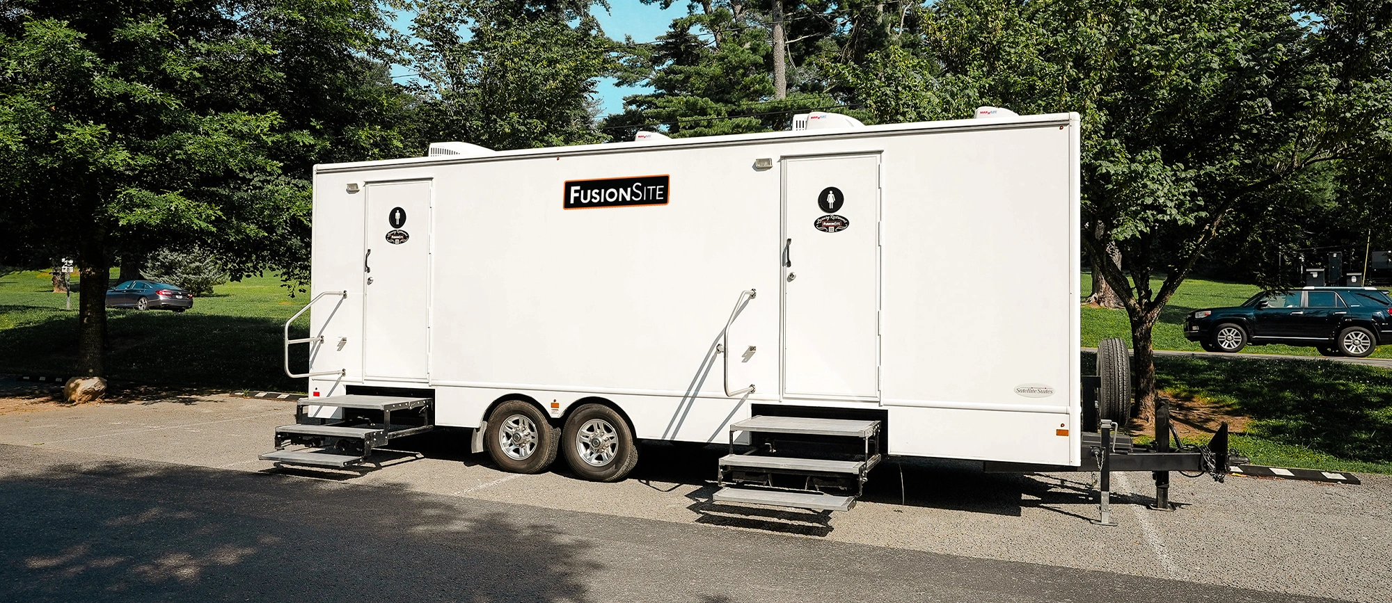 A large white portable restroom trailer with two entry doors is parked on a paved lot surrounded by trees, with a car visible in the background on a sunny day.