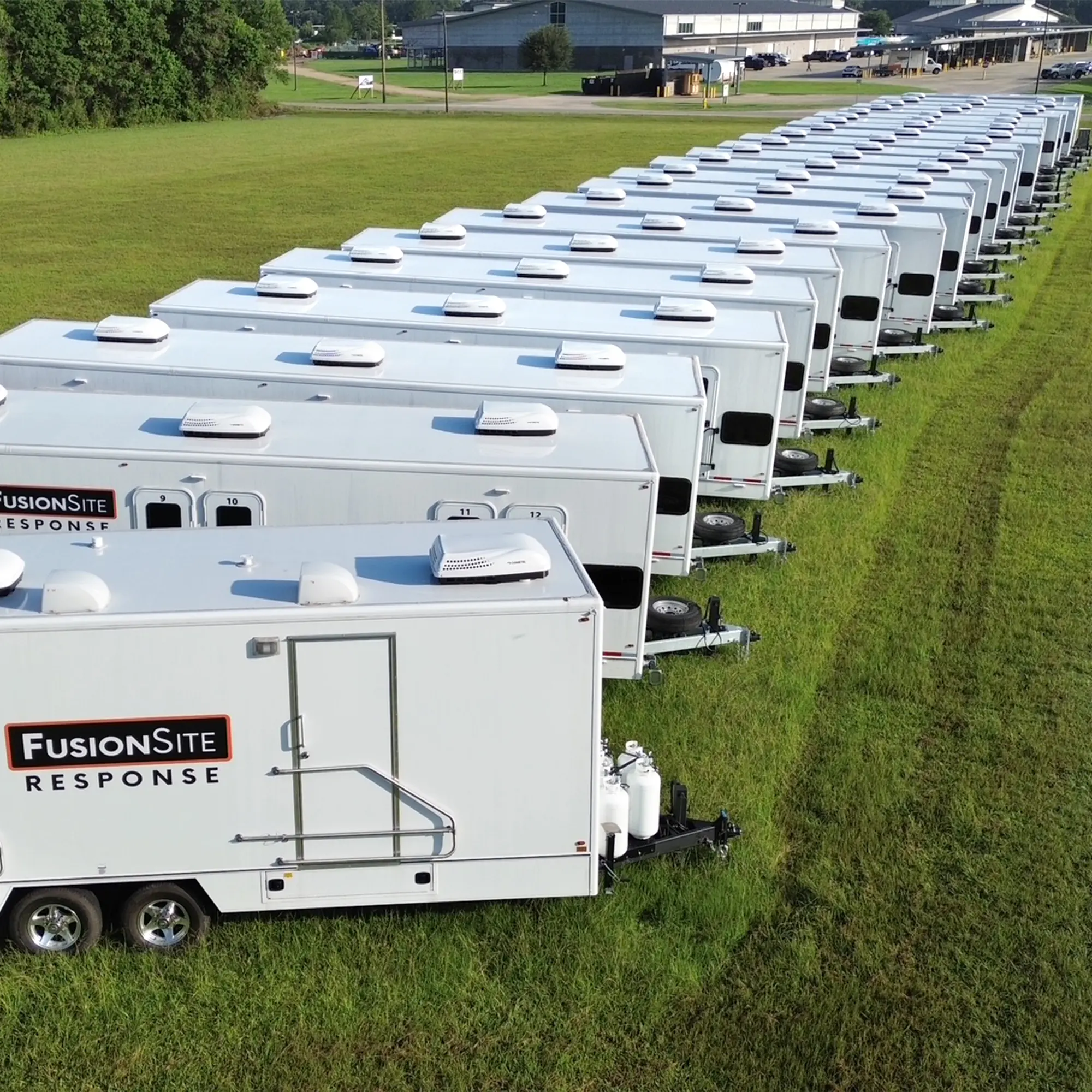 A row of white portable restroom trailers is parked on a grassy field, with a tree line in the background and a building visible in the distance under a clear blue sky.