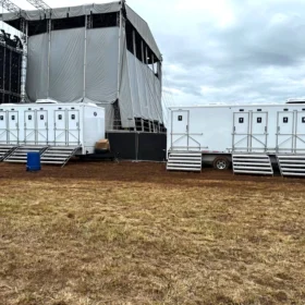A row of white portable restroom trailers is positioned on a muddy field next to a large stage structure, with overcast skies overhead.