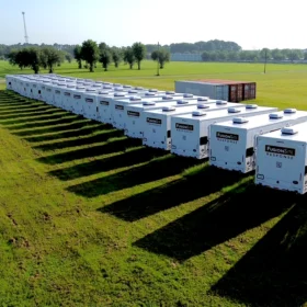 A row of white portable restroom units is lined up on a grassy field, casting long shadows under the morning sun, with a storage container visible in the background.