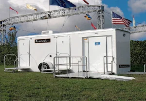 A white portable restroom trailer with wheelchair access is positioned on a grassy field, surrounded by trees and under a cloudy sky, with colorful flags displayed on a nearby structure.
