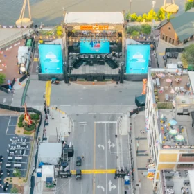 A row of portable restrooms is set up along a busy street near outdoor seating areas and a stage, with buildings and vehicles visible in the surrounding urban environment.