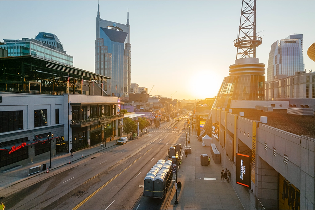 A row of portable restrooms is positioned along a city street near a large building, with the sun rising in the background and pedestrians walking by on a clear morning.