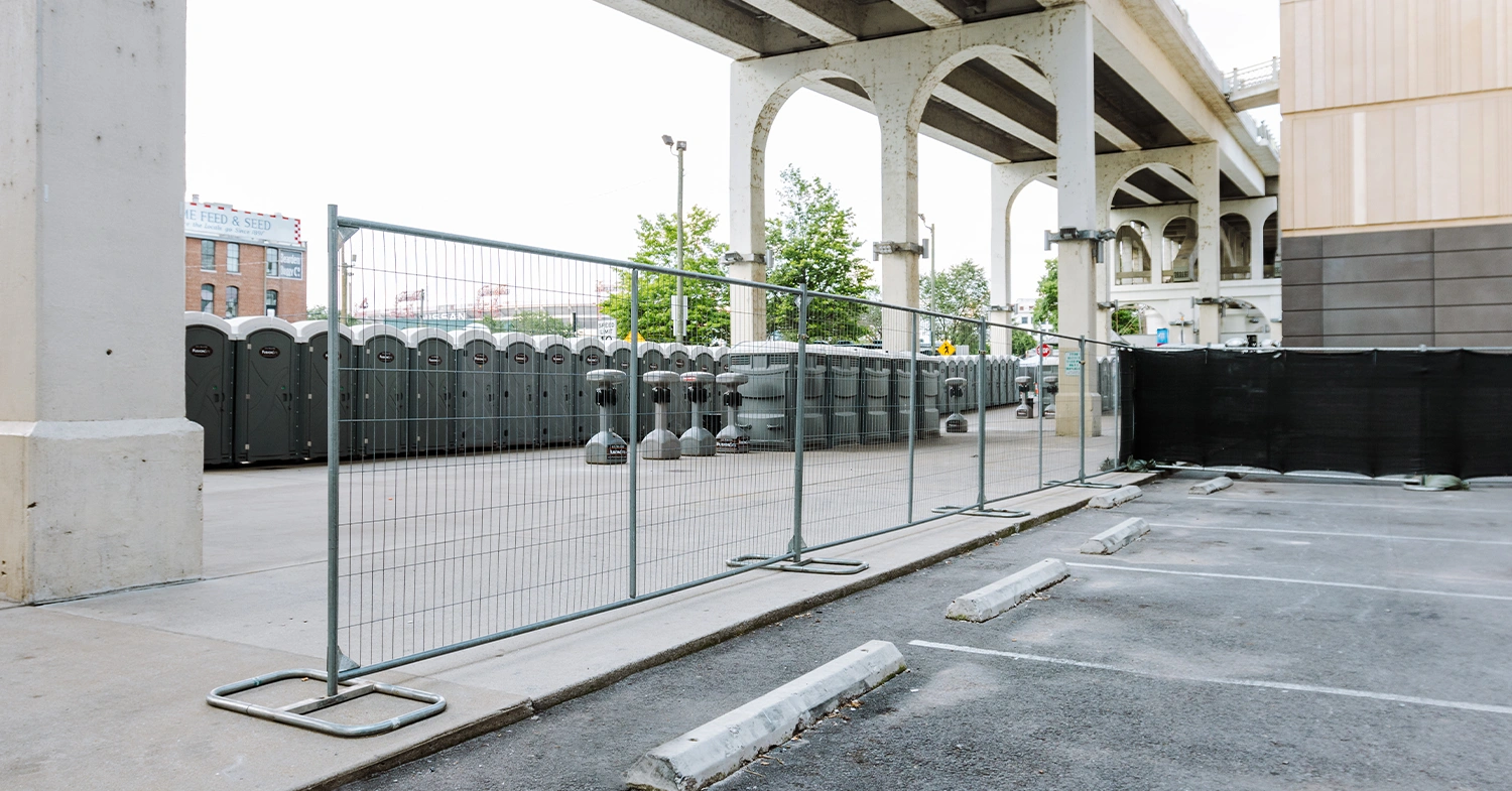A row of portable restrooms is lined up behind a construction fence in a parking lot, with a concrete overpass and greenery visible in the background on a cloudy day.