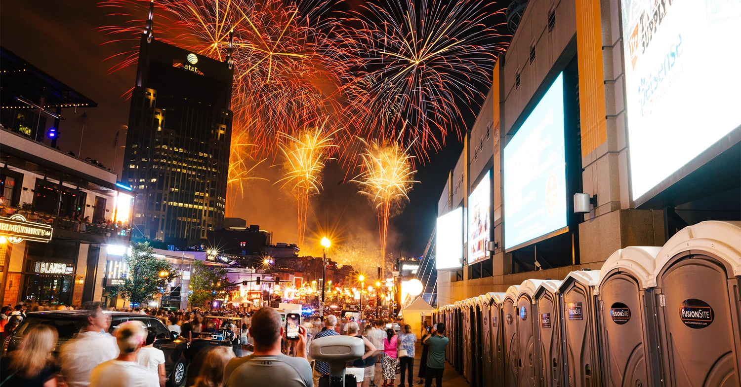 A row of portable restrooms is positioned along a bustling street, illuminated by colorful fireworks in the night sky, with a crowd of people celebrating nearby.