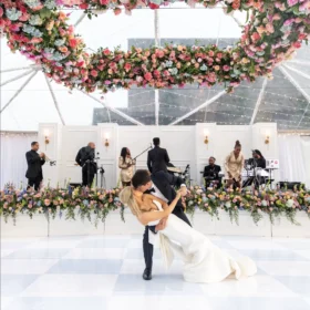 A row of elegant portable restrooms is set up beside a beautifully decorated outdoor wedding venue, featuring a floral arch and a live band performing under a clear tent on a bright day.