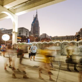 A row of portable restrooms is positioned along a concrete walkway under a large overpass, with a bustling crowd of people moving past in the background as evening sets in.