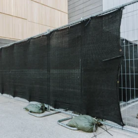 A temporary metal fence covered with black privacy mesh and secured with sandbags at the base along a modern building exterior.