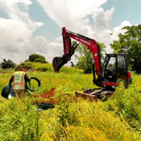 A red excavator is digging in a grassy field surrounded by tall weeds, while a worker in a safety vest carries a circular plastic component under a partly cloudy sky.