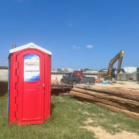 A red portable restroom is positioned on a grassy area next to a concrete wall, with a construction site visible in the background featuring heavy machinery and stacked logs under a clear blue sky.